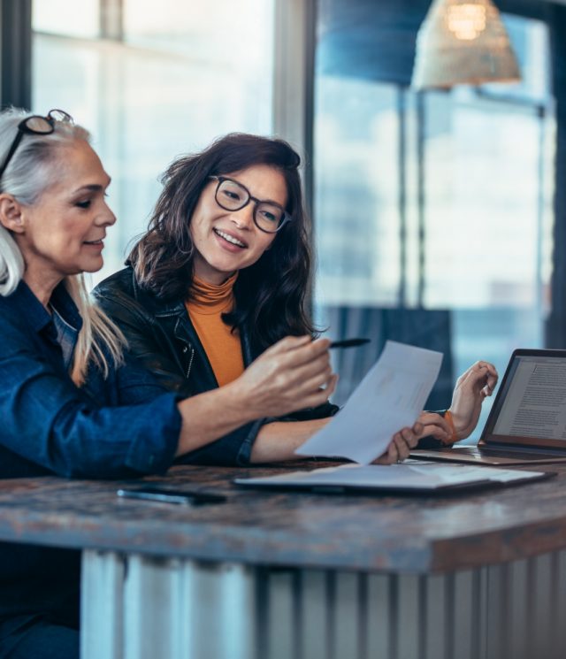 Two women analyzing documents while sitting on a table in office. Woman executives at work in office discussing some paperwork.