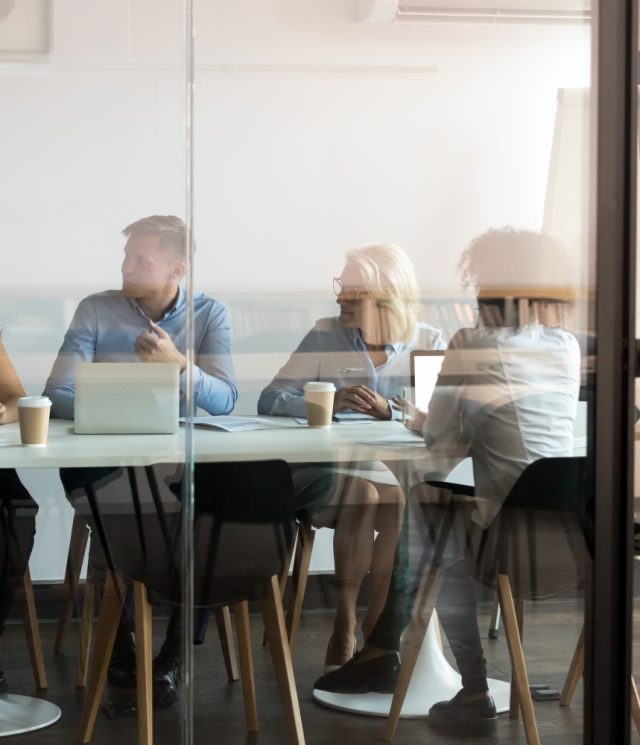 Advertising department brainstorming at modern office boardroom behind closed doors, view through the glass wall. Diverse staff led by ceo discussing new project sharing ideas thoughts and sales pitch