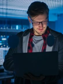 Portrait of a Smart Focused Young Man Wearing Glasses Holds Laptop. In the Background Technical Department Office with Specialists Working and Functional Data Server Racks