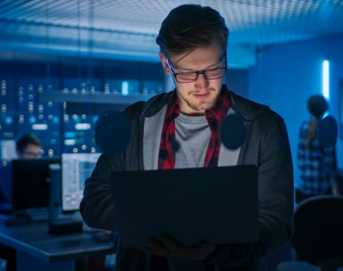 Portrait of a Smart Focused Young Man Wearing Glasses Holds Laptop. In the Background Technical Department Office with Specialists Working and Functional Data Server Racks