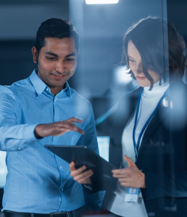 In Technology Research Facility: Female Project Manager Talks With Chief Engineer, they Consult Tablet Computer. Team of Industrial Engineers, Developers Work on Engine Design Using Computers